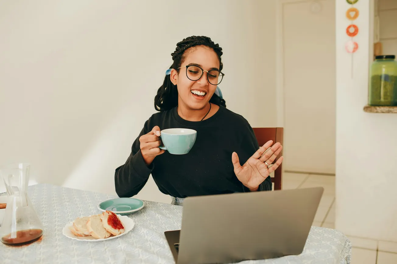 A woman with glasses smiles while holding a teal mug, engaged in a video call on a laptop. A plate of pastries is on the table.
