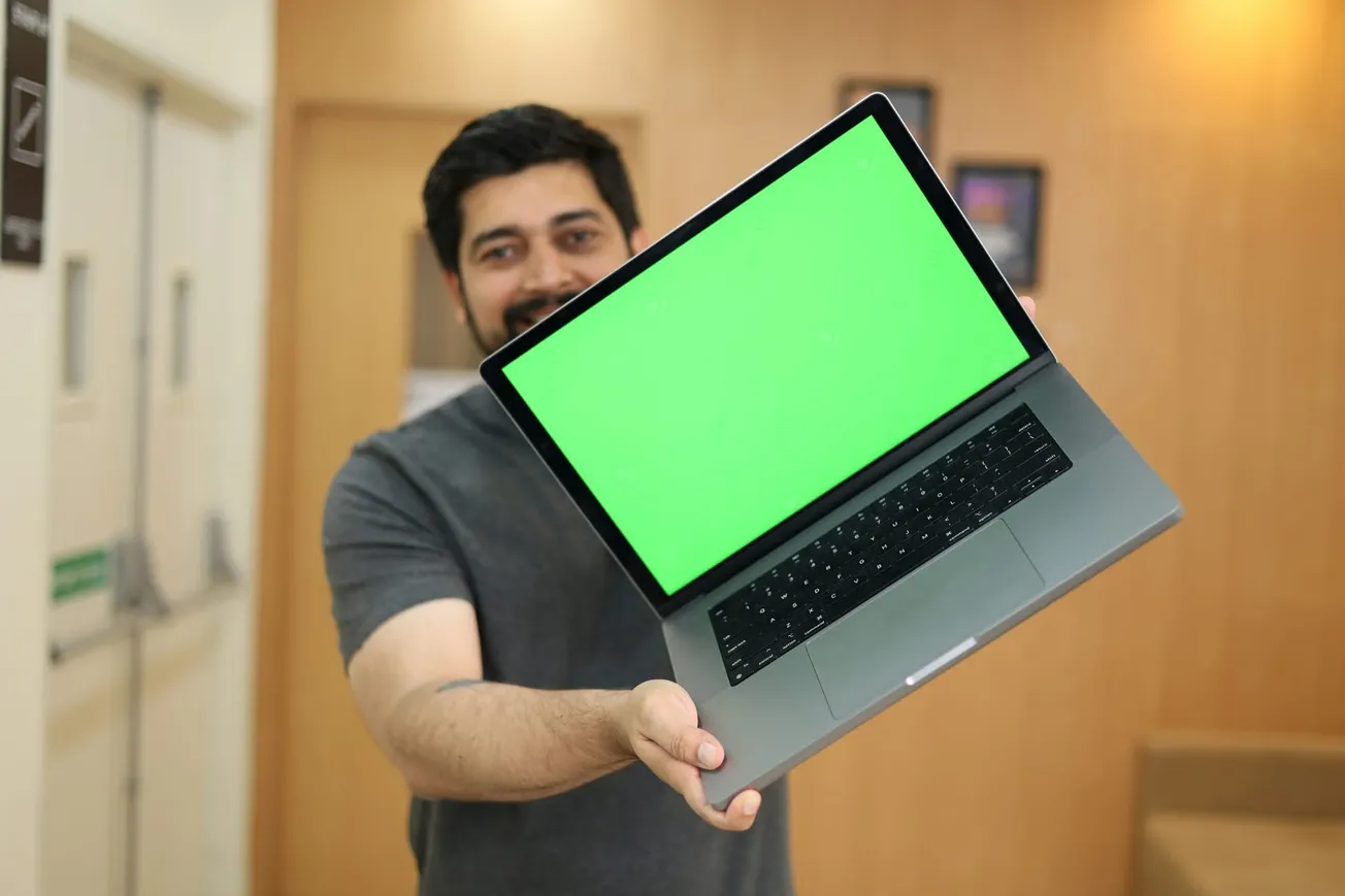 Man holding a laptop with a bright green screen, standing in a room with beige walls. The setting is casual and informal, conveying a sense of approachability.