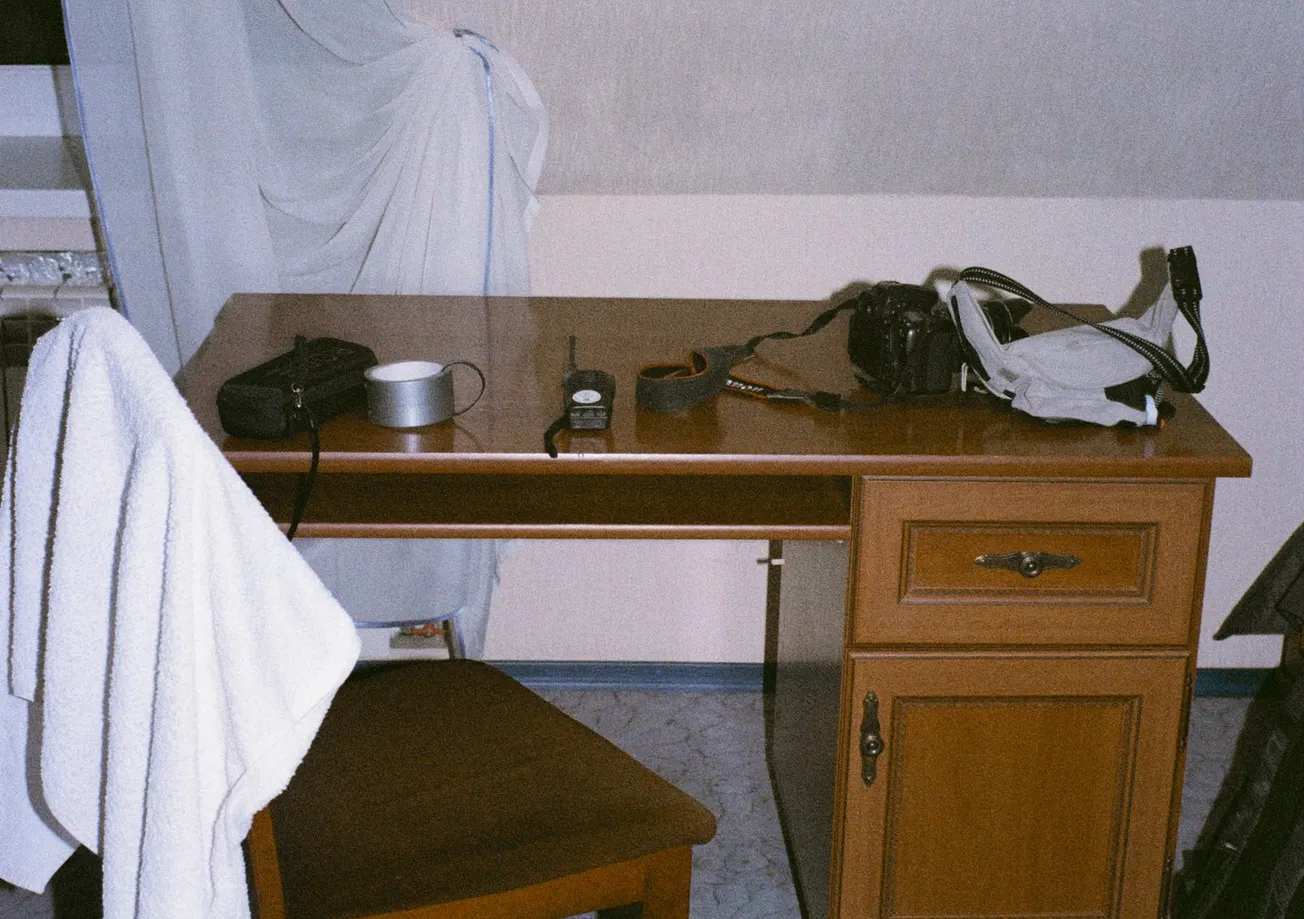 A wooden desk with items like a camera, a face mask, tape, a radio, and a towel-draped chair. A curtain hangs in the background, suggesting a casual setting.