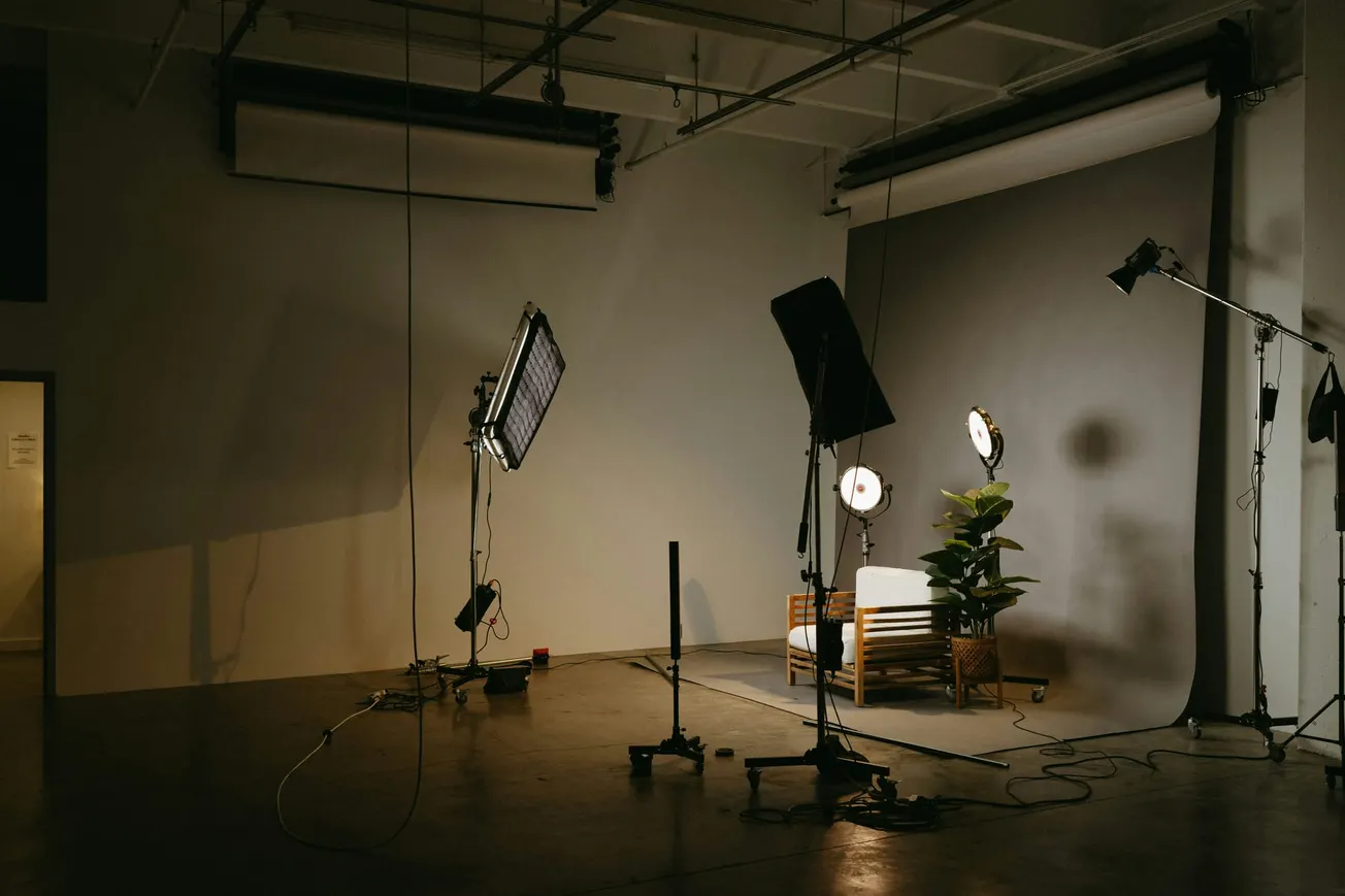 A dimly lit photo studio with lighting equipment surrounding a wooden chair on a white cushion. A potted plant adds a touch of greenery on a gray backdrop.
