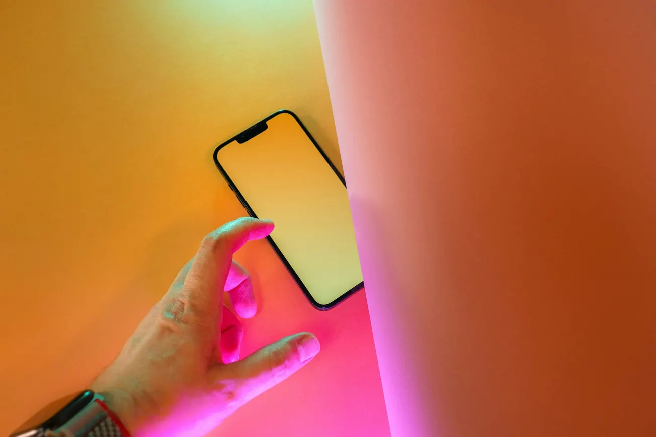 A hand reaches for a smartphone on a vibrant yellow and pink background. The scene is illuminated by colorful neon lights, creating a modern, tech-savvy vibe.