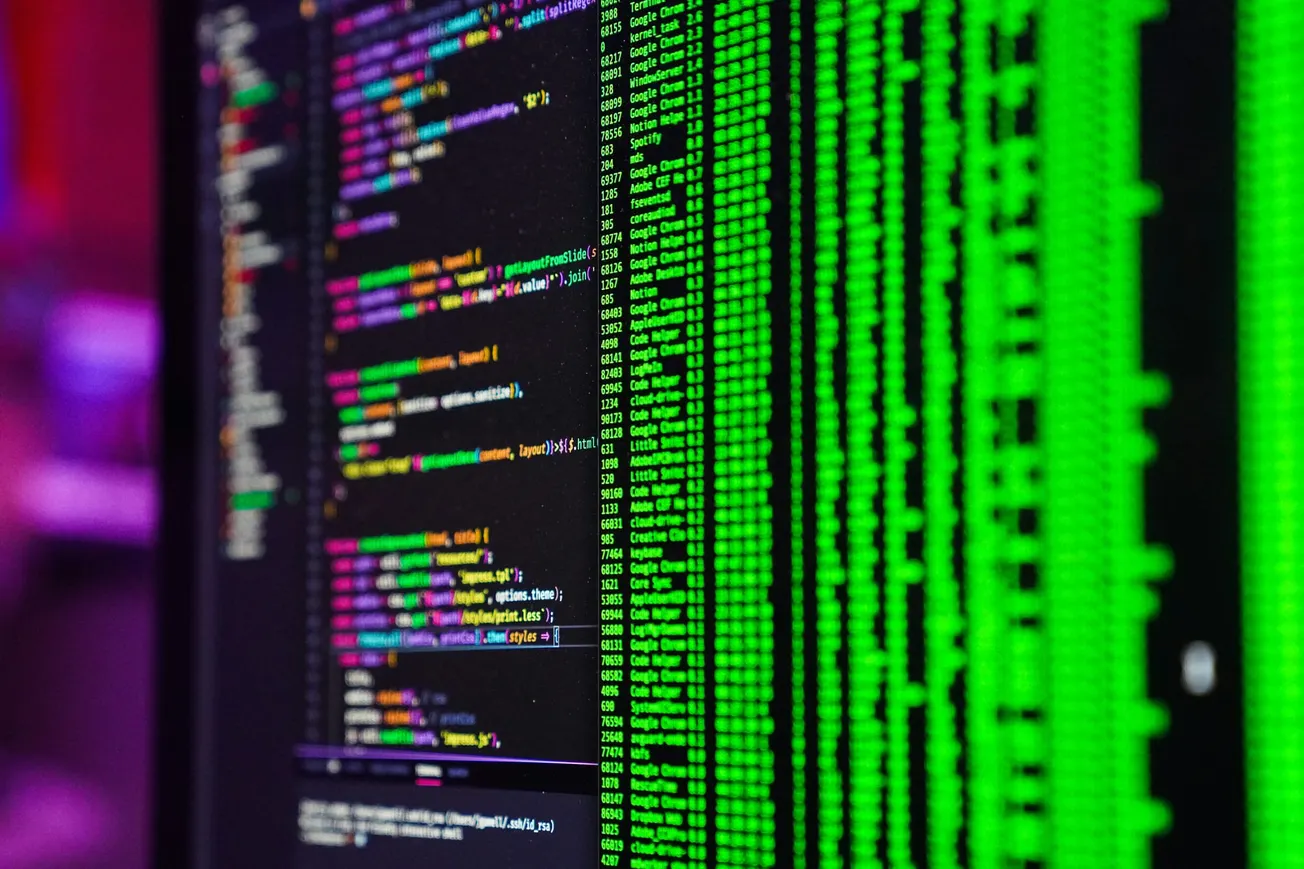 Close-up of computer screens displaying colorful code and green text, creating a high-tech, focused atmosphere. Lines of code suggest programming work.