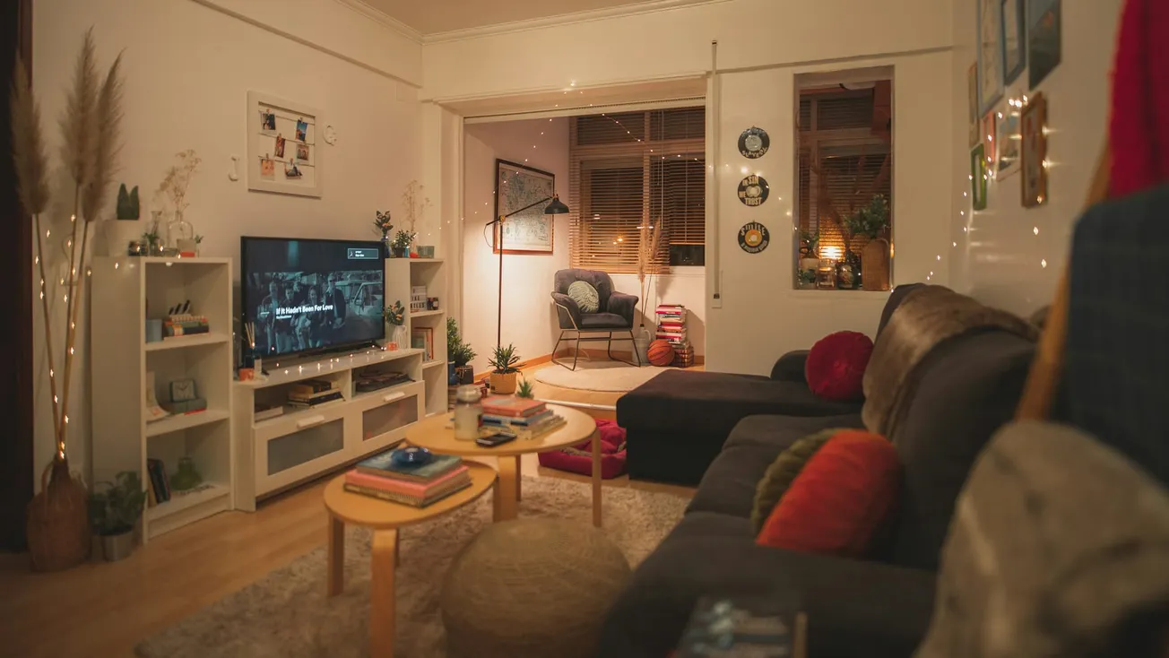 Cozy living room with dim lighting, featuring a large sofa with colorful cushions, a TV on a shelf, a small coffee table, and warm decor elements.