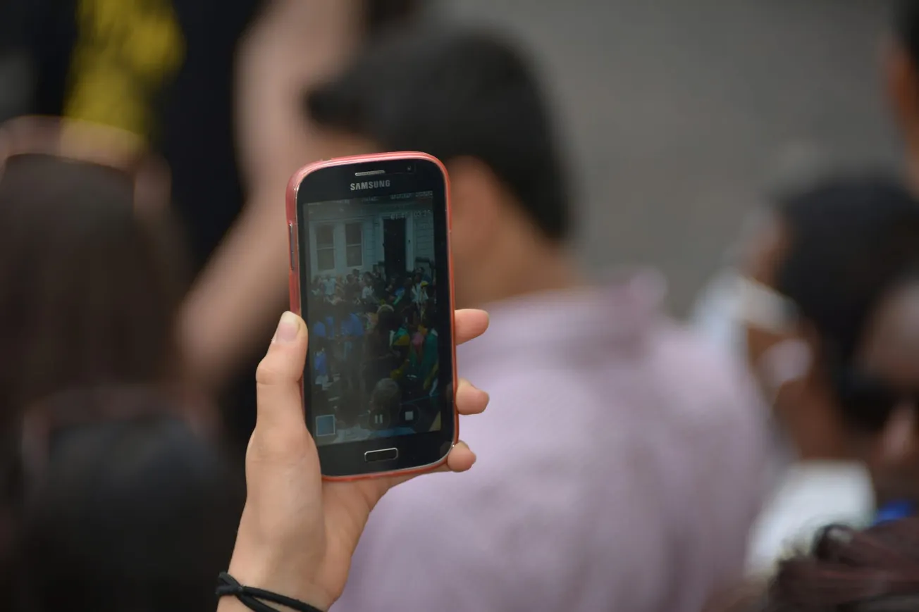 A hand holding a Samsung phone captures a crowded event. The phone screen shows people seated outside a building, conveying a lively gathering.