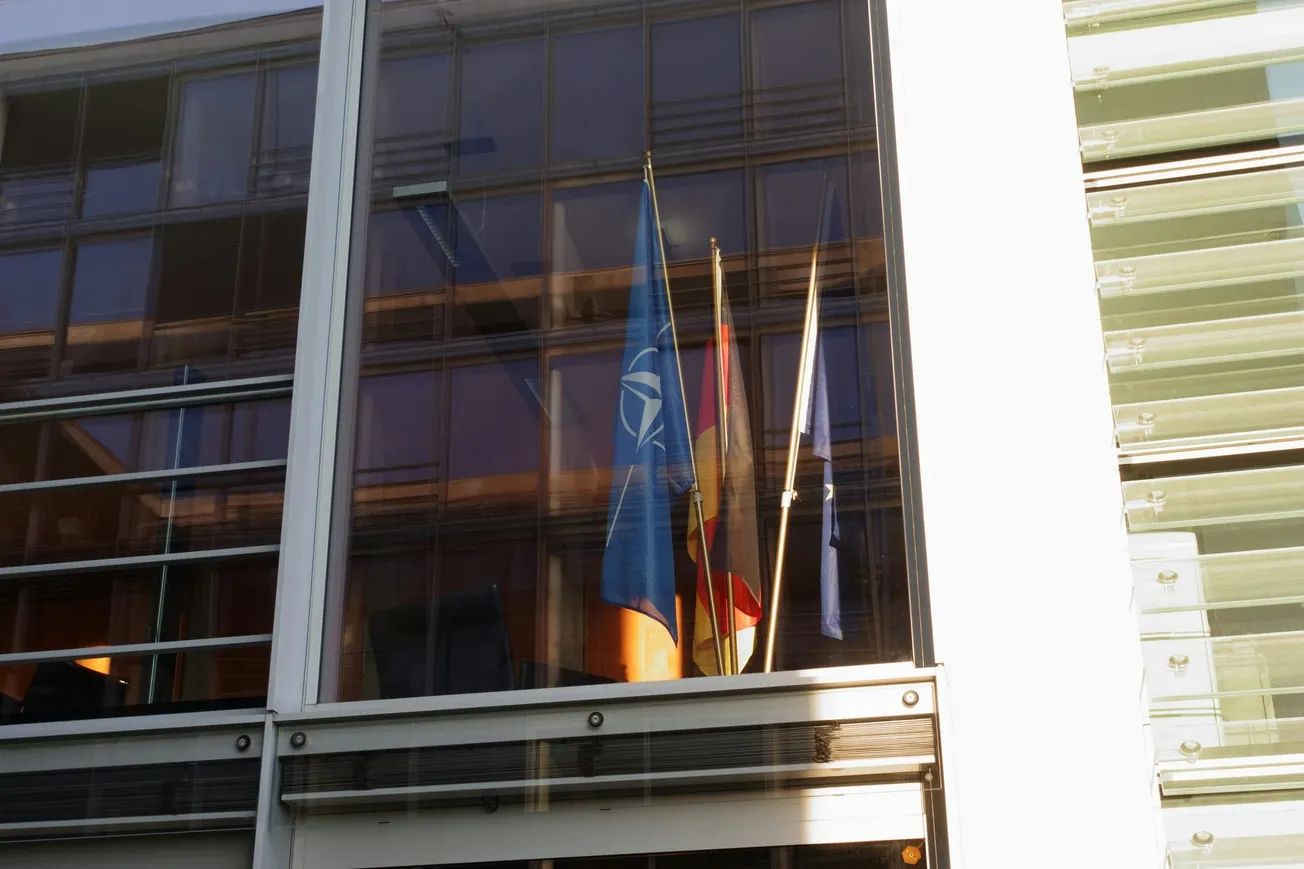 Flags, including a blue one with a white emblem, are seen through a large window reflecting light on a modern building's facade.