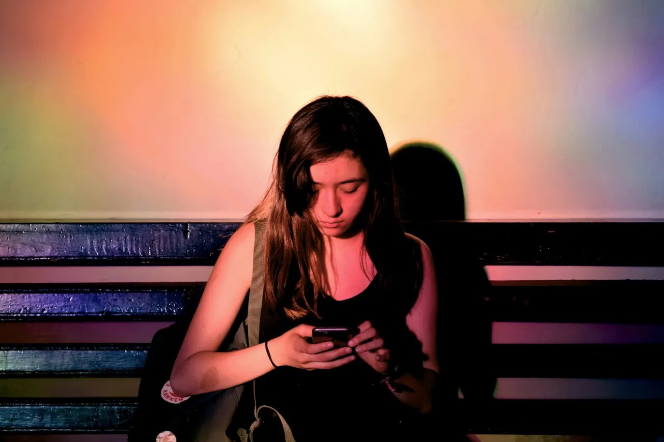 A young woman sits on a bench, focused on her phone. Multicolored lights cast a warm, serene glow, creating a contemplative and peaceful atmosphere.