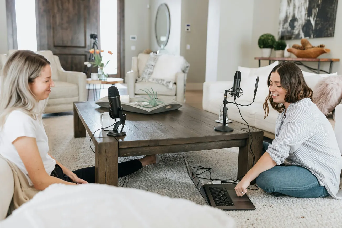 Two women sit on the floor in a cozy living room, recording a podcast. They smile at each other with microphones on a wooden table between them.