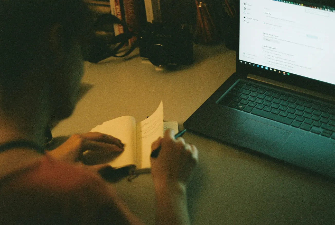 A person writes in a notebook at a desk beside an open laptop displaying a website. The setting is dimly lit, creating a focused, studious atmosphere.