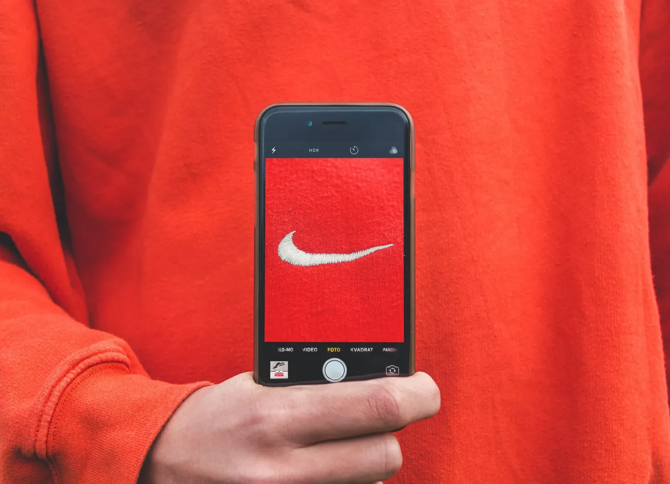 A person in an orange sweatshirt holds a phone displaying a close-up of a white Nike logo on red fabric, emphasizing a sporty, modern vibe.
