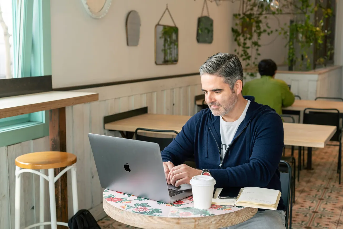 Man with gray hair works on a laptop at a cafe, surrounded by greenery. He looks focused, with a coffee cup and notebook beside him on the table.