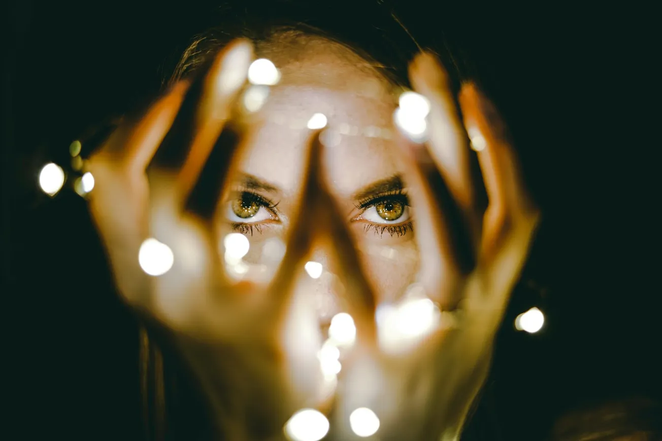 A person's intense eyes peer through their hands, which cradle glowing string lights in a dark setting, creating a mysterious and enchanting atmosphere.