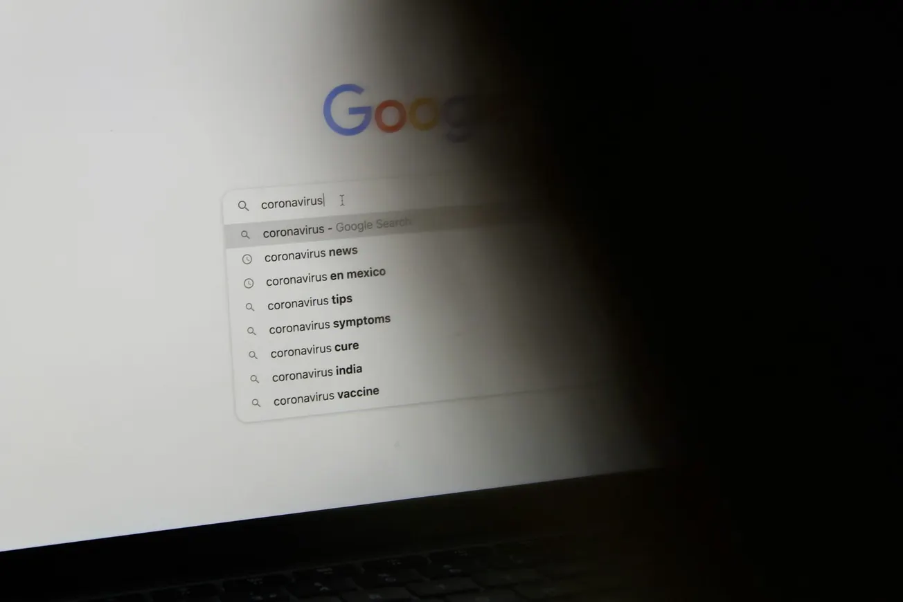A computer screen showing a Google search for "coronavirus" with suggestions like news, symptoms, and vaccine. Dark shadow partially obscures the screen.