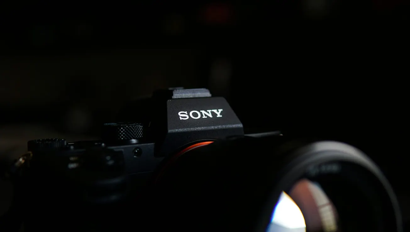 Close-up of a Sony camera in a dark setting, highlighting the brand logo. The lens reflects a soft light, creating a focused and professional tone.
