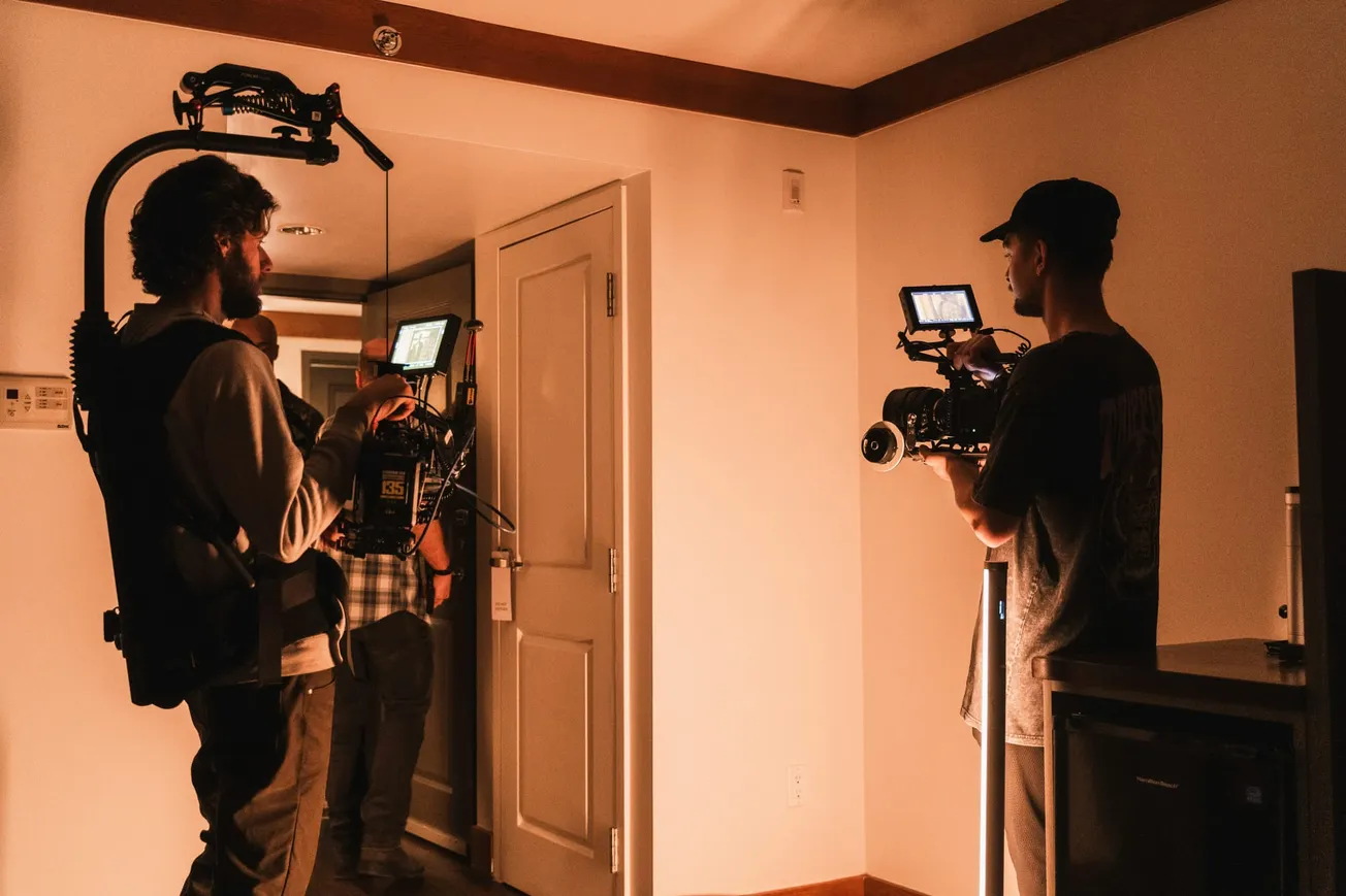 Two videographers with cameras, one in a harness, film inside a dimly lit room. A third person is partially visible in a doorway, setting a behind-the-scenes mood.