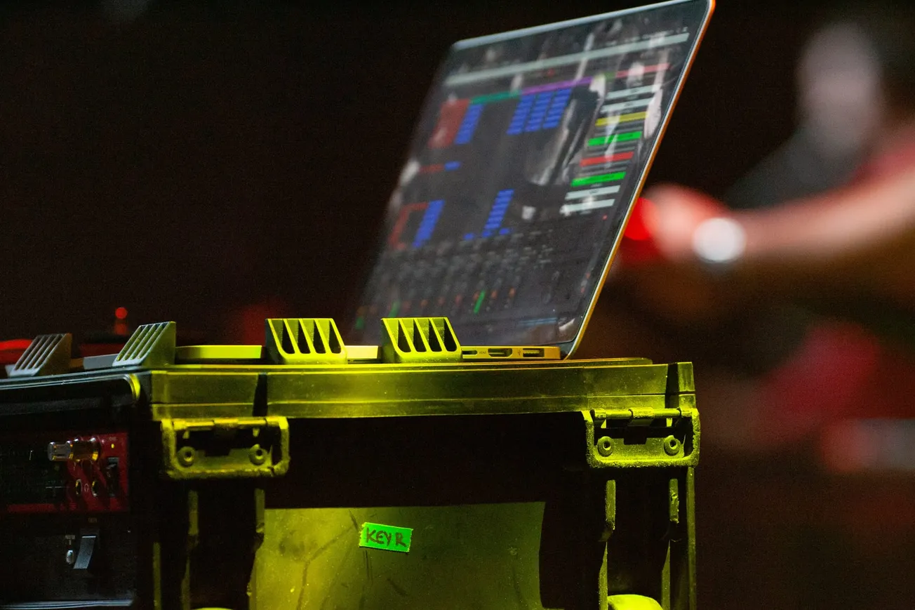 A laptop displaying colorful audio software on its screen sits atop a rugged equipment case on stage, with a dimly lit background suggesting a concert setting.