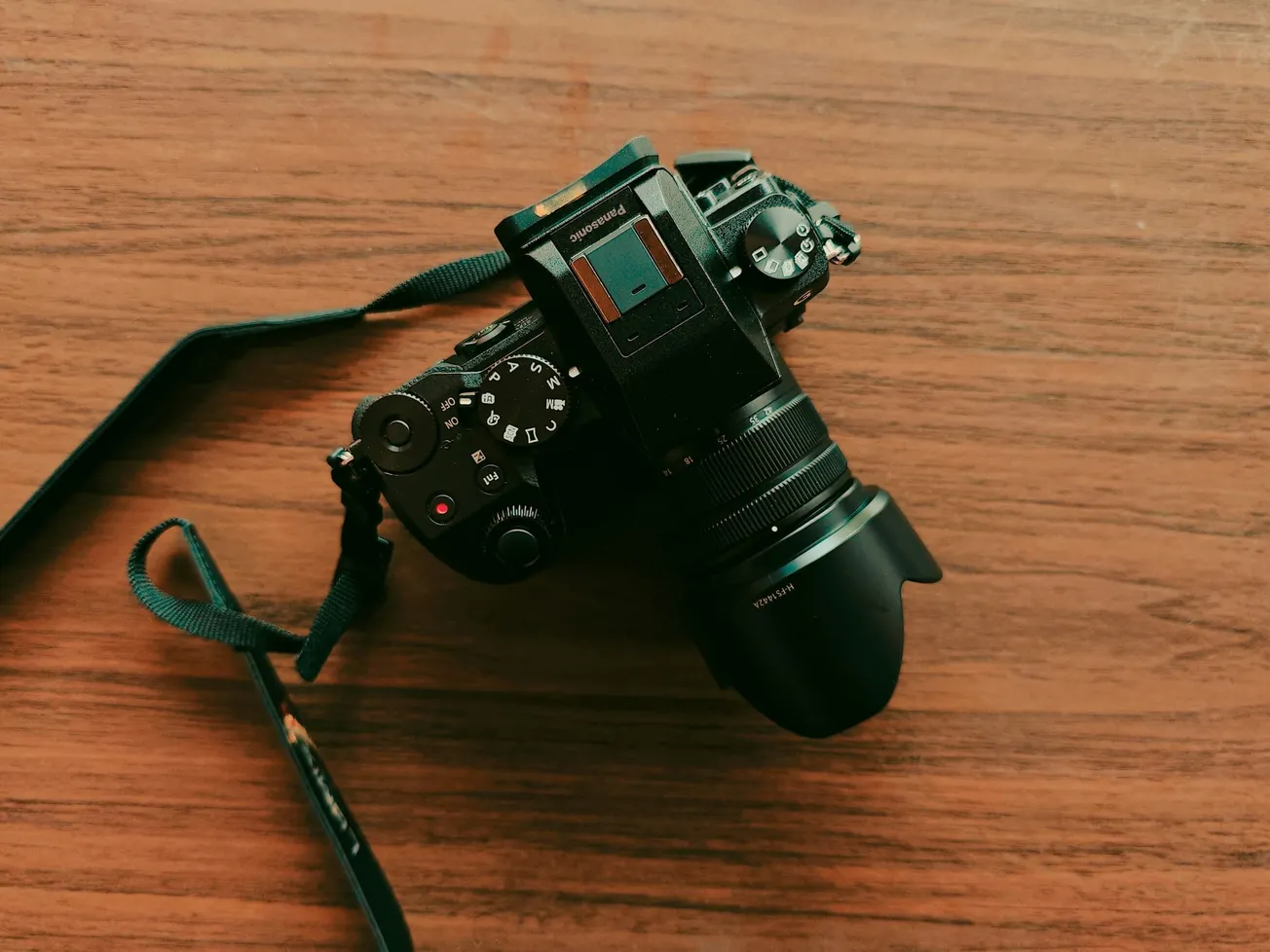 A black camera with a lens cap rests on a wooden table. The camera strap is loosely attached, and the lighting creates a warm, vintage tone.