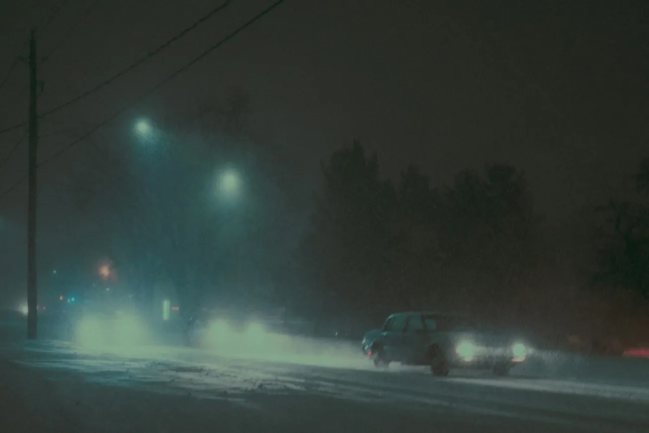 Night scene with a vintage car driving on a snowy road. Streetlights cast a hazy glow over the scene, creating a mysterious, moody atmosphere.