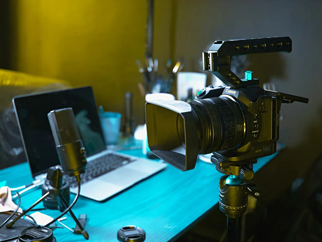 A professional camera on a tripod faces a desk with a laptop and microphone, under soft lighting. The scene conveys a creative, tech-focused workspace.