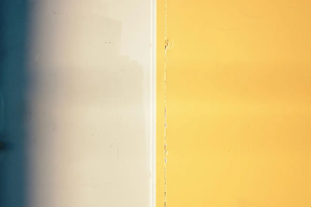 Split image of a wall, left side in shadow blue, right side bright yellow. Light and shadow create a stark, modern contrast, evoking simplicity and calm.