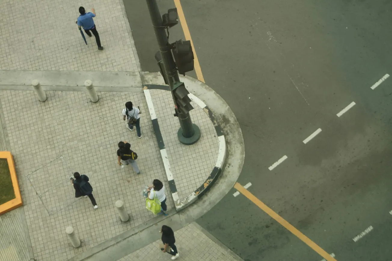 Aerial view of pedestrians crossing a tiled sidewalk near a curved street corner with a traffic light. The scene is casual and urban, with muted colors.