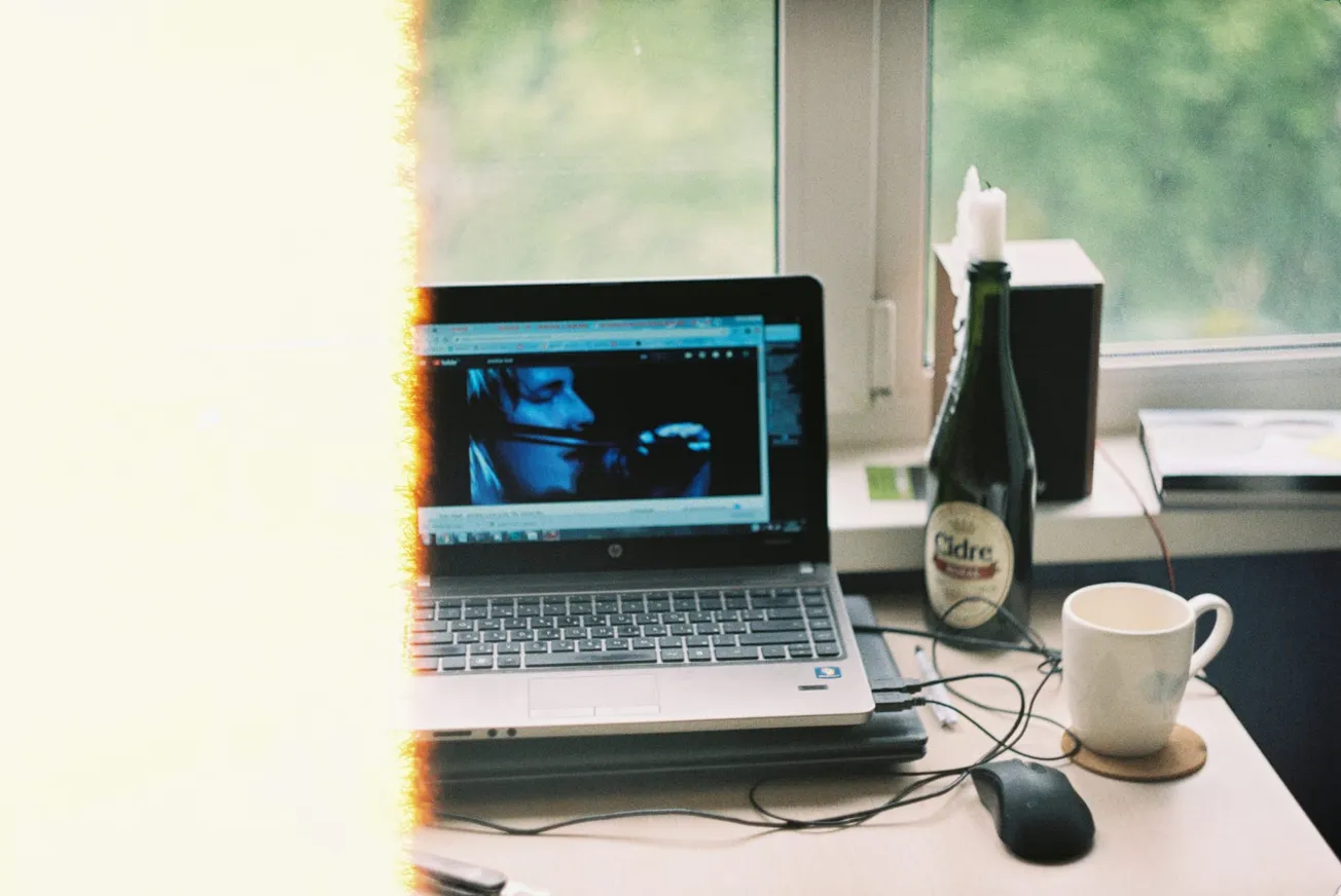 A laptop on a desk displays a video; a light leak obscures part of the image. Nearby are a cup, mouse, cider bottle, and a window with greenery outside.