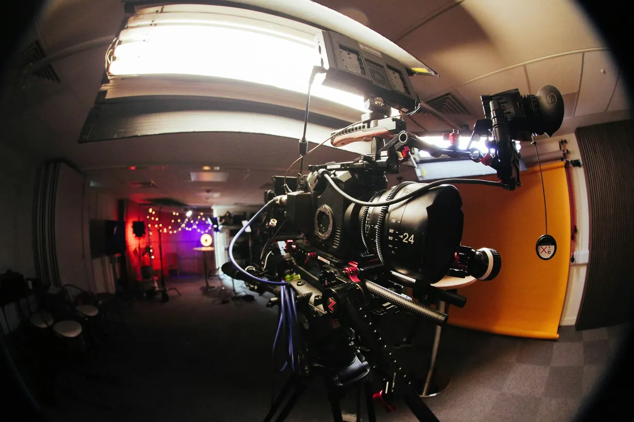 Close-up of a professional film camera on a set, surrounded by lights and colorful backdrops. The scene has a creative, bustling atmosphere.