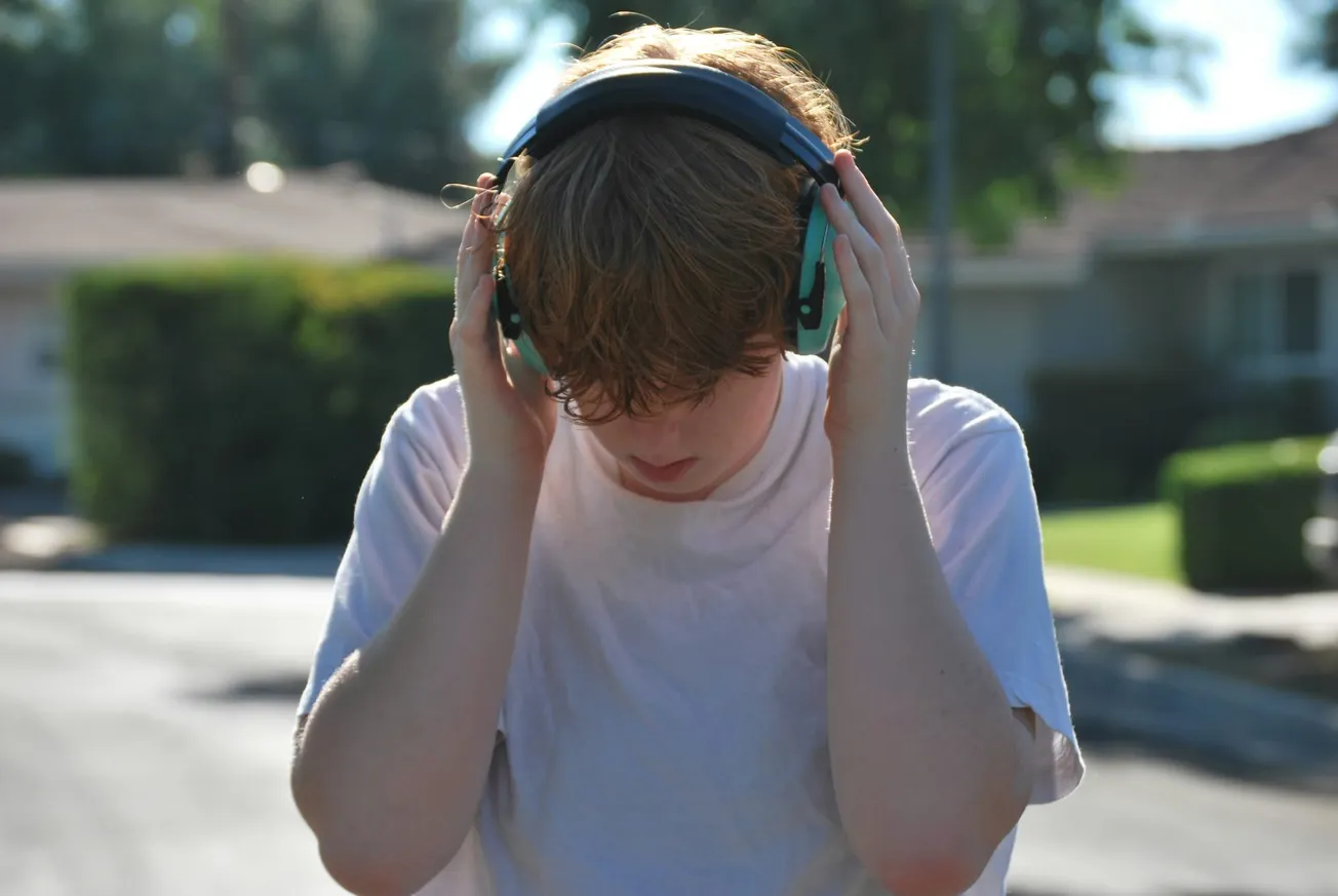 A person with brown hair and a white shirt holds large headphones over their ears, standing outdoors in sunlight with houses and greenery in the background.