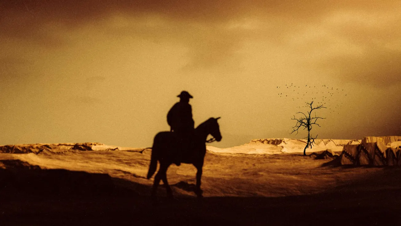 Silhouette of a lone rider on horseback in a barren desert landscape during sunset. A solitary leafless tree stands in the distance under a dramatic sky.