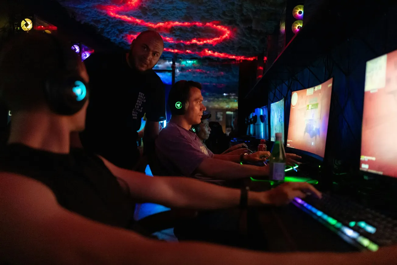 In a dimly lit gaming room, three people wearing headphones focus intently on their glowing screens. The ceiling displays vivid red and blue lights, adding a vibrant atmosphere.