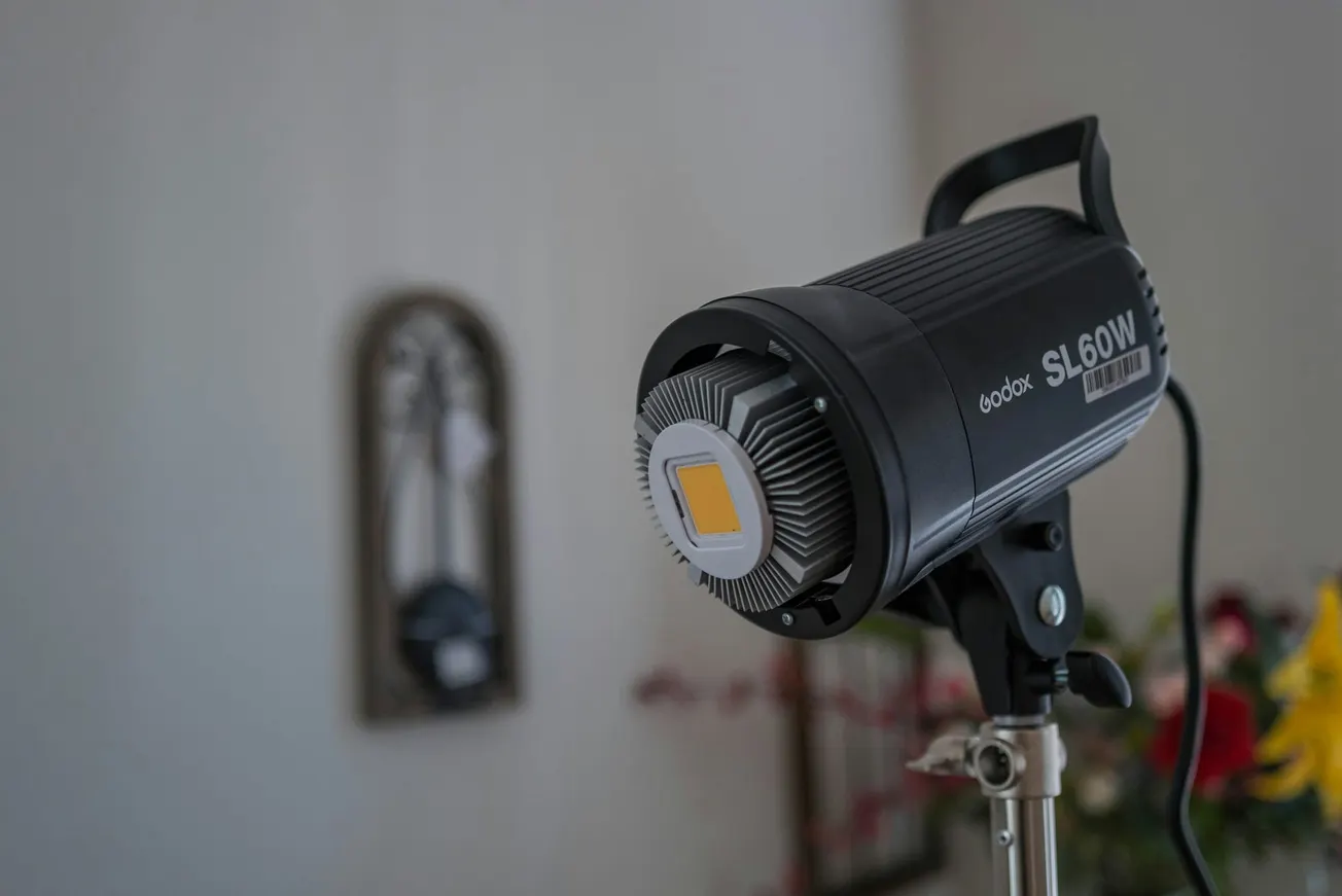 Close-up of a Godox SL60W studio light on a stand in a softly lit room. In the background, there's a blurred wall decor and colorful flowers.
