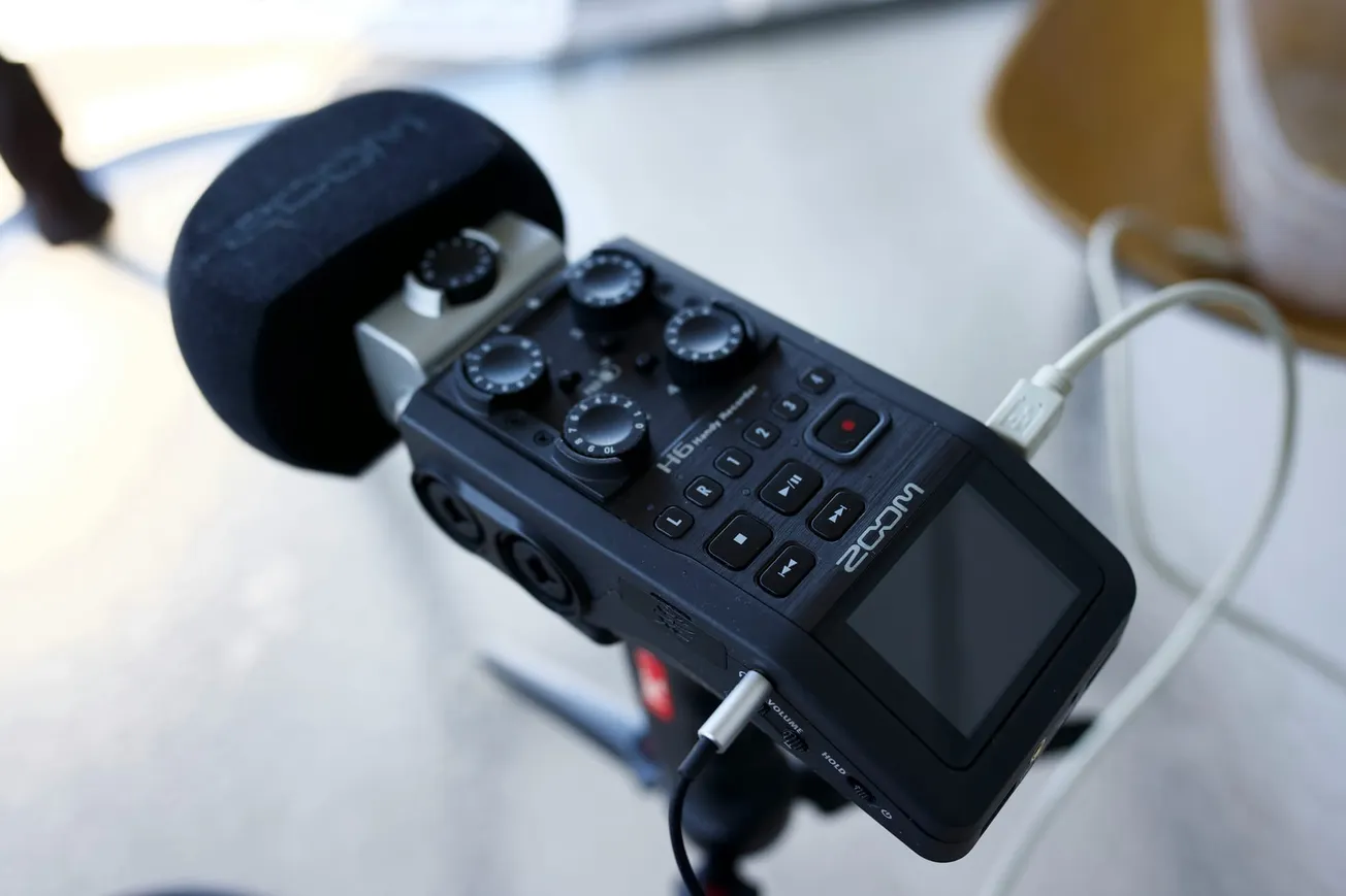Close-up of a Zoom audio recorder with a microphone attached, featuring multiple control knobs and buttons. It rests on a tripod, evoking a professional setting.