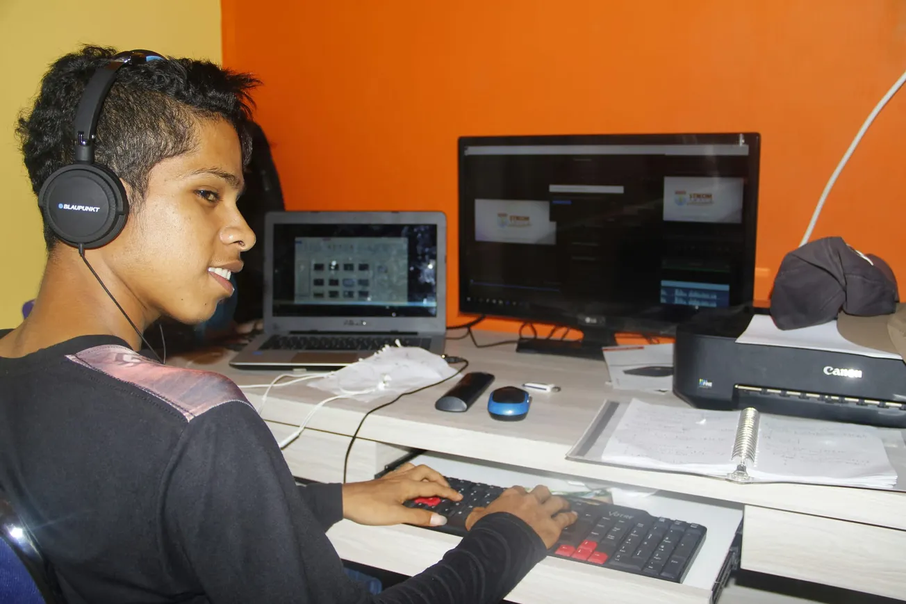 A person wearing headphones works at a desk with a laptop, a monitor, and a keyboard. The room features orange walls, creating a focused atmosphere.