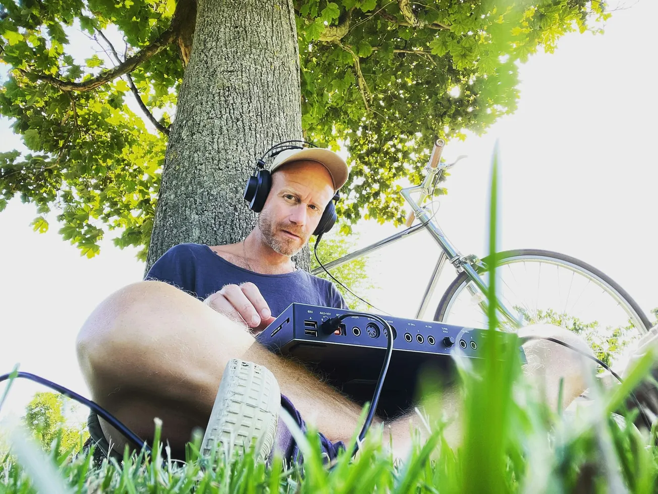 Man in headphones sits under a tree, intently using a musical device. A bicycle leans nearby. The atmosphere is relaxed and creative.