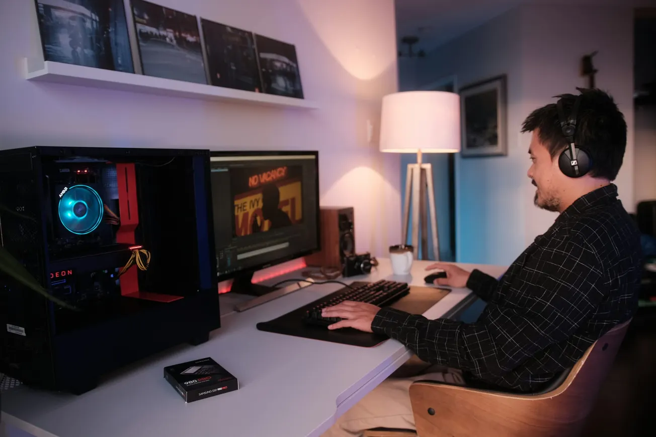 A man video editing on a professional PC gaming setup with RGB lighting, dual monitors, and a Samsung 980 PRO SSD on the desk.
