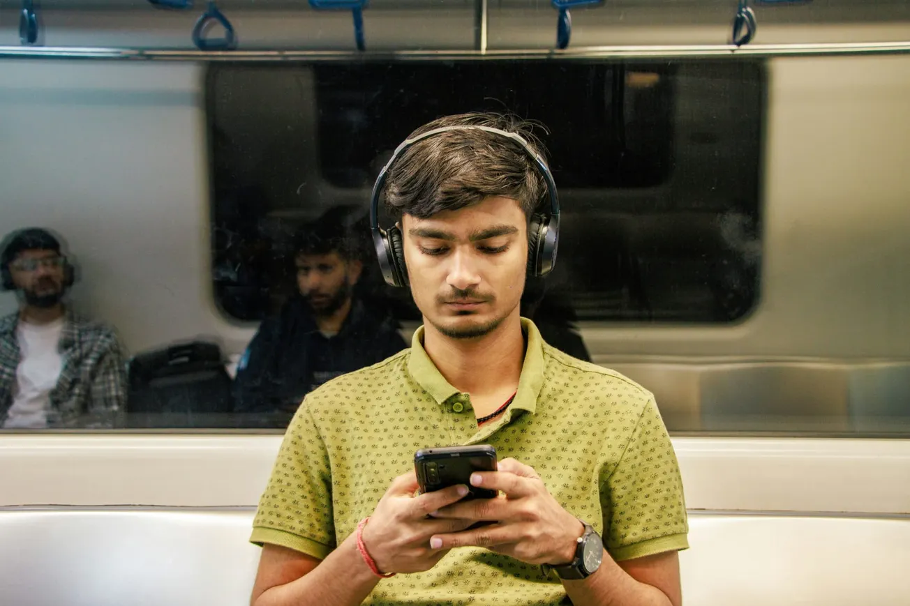 A young man with headphones, in a green shirt, focused on his phone while sitting on a train. Reflections show other passengers, creating a calm mood.