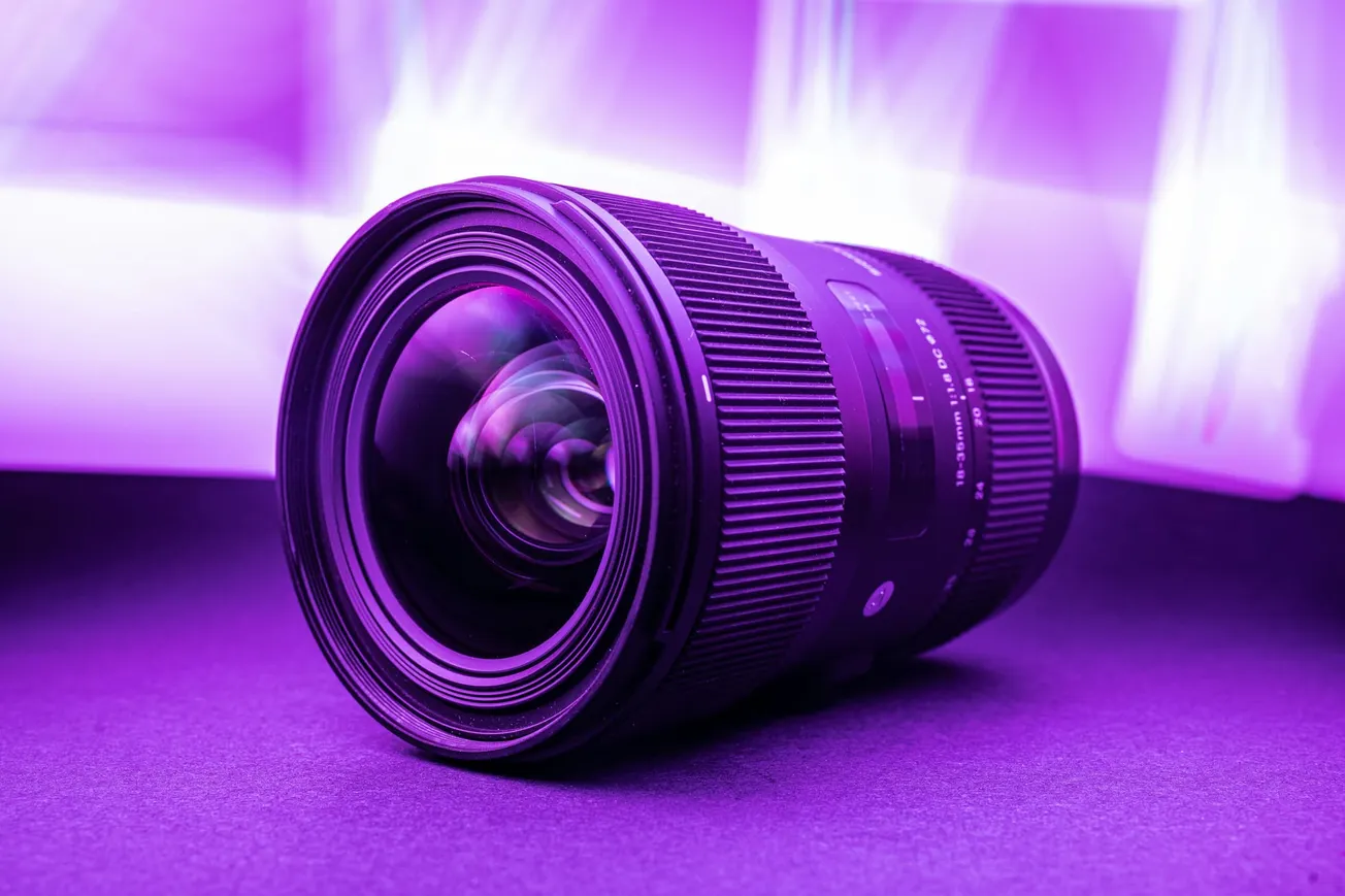 A camera lens lies on its side, surrounded by a vibrant purple light. The background is blurred, creating a modern and dynamic atmosphere.