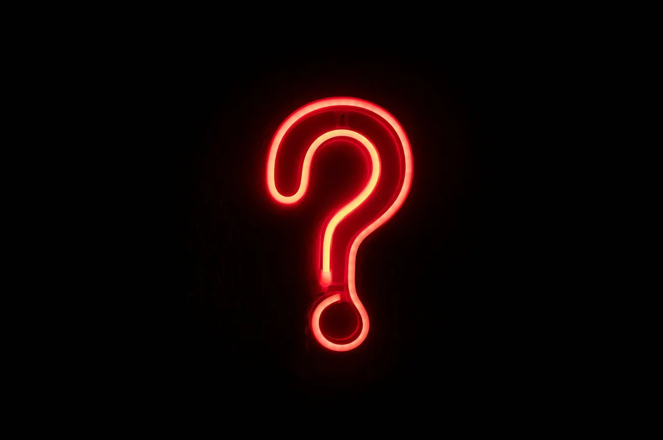 A glowing red neon question mark on a black background, conveying a sense of mystery and curiosity. The vibrant light contrasts sharply with the darkness.