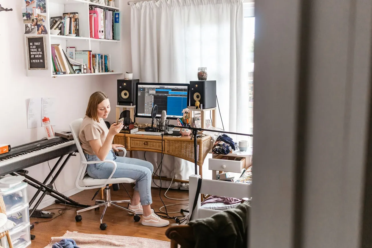 A woman sits at a desk with a computer and audio setup, checking her phone. A keyboard and shelves with books are visible, conveying a creative workspace.