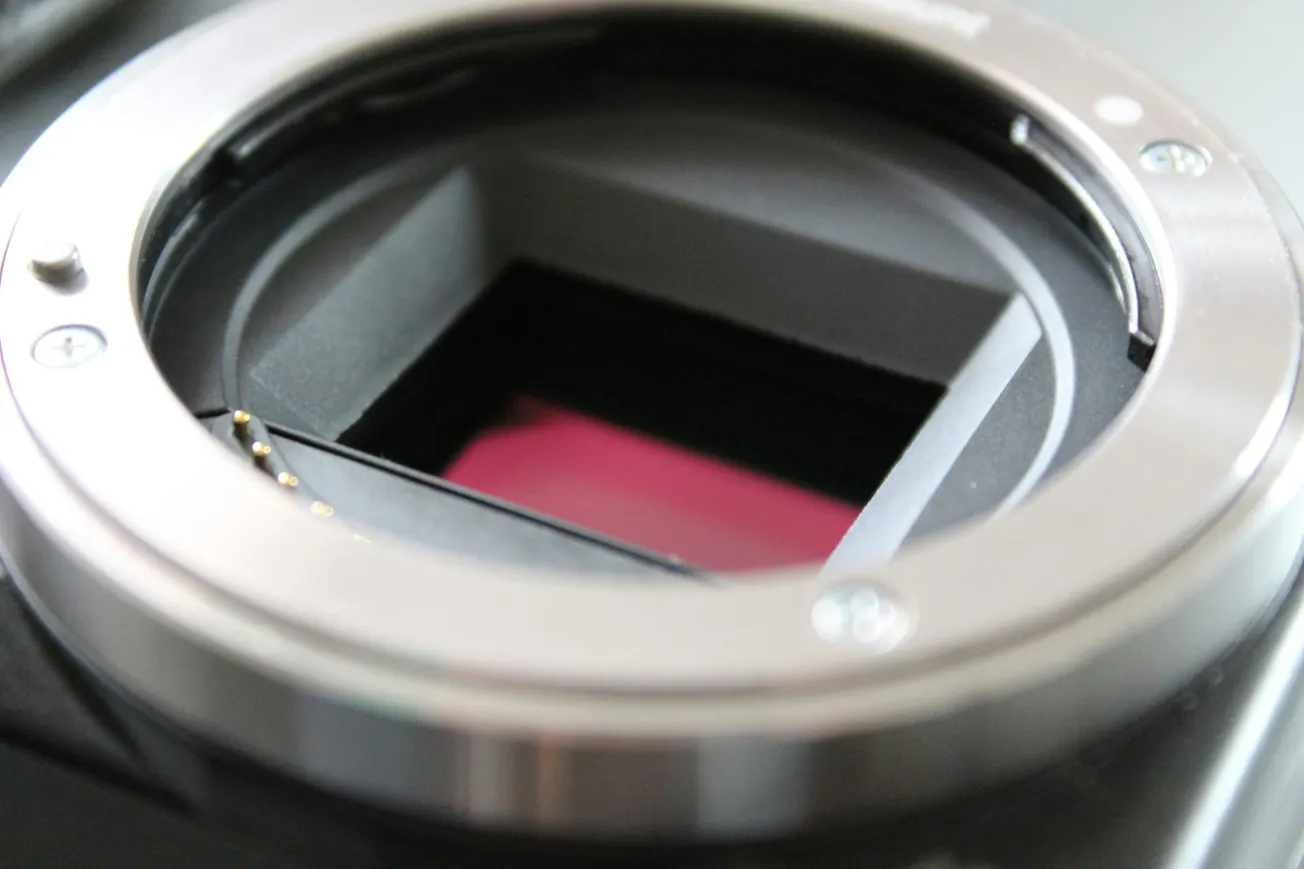 Close-up of a camera's lens mount showing the sensor inside. The metal edges contrast with the pinkish hue of the sensor, evoking a sense of precision.