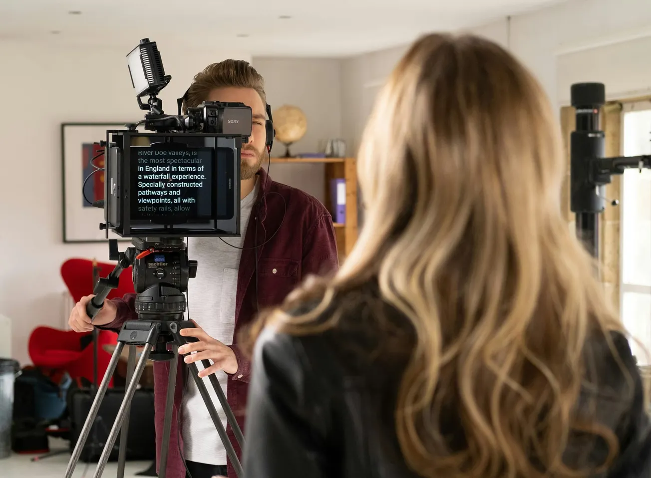 A camera operator films a woman indoors. The operator wears headphones, the camera has a teleprompter, and a microphone is visible. The scene feels professional.