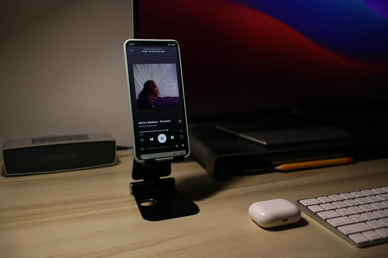 A smartphone on a stand plays music on a desk. Nearby are a keyboard, AirPods, speaker, and a large screen with a colorful background, creating a tech-savvy ambiance.