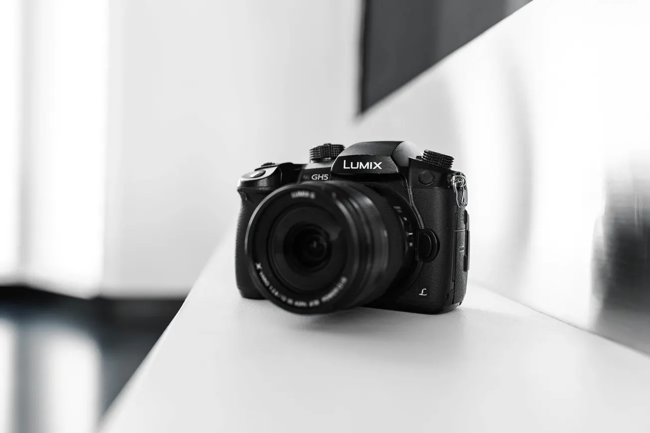 A black Panasonic Lumix GH5 camera sits on a sleek, white surface, its lens focused forward. The background is softly blurred, emphasizing the camera.