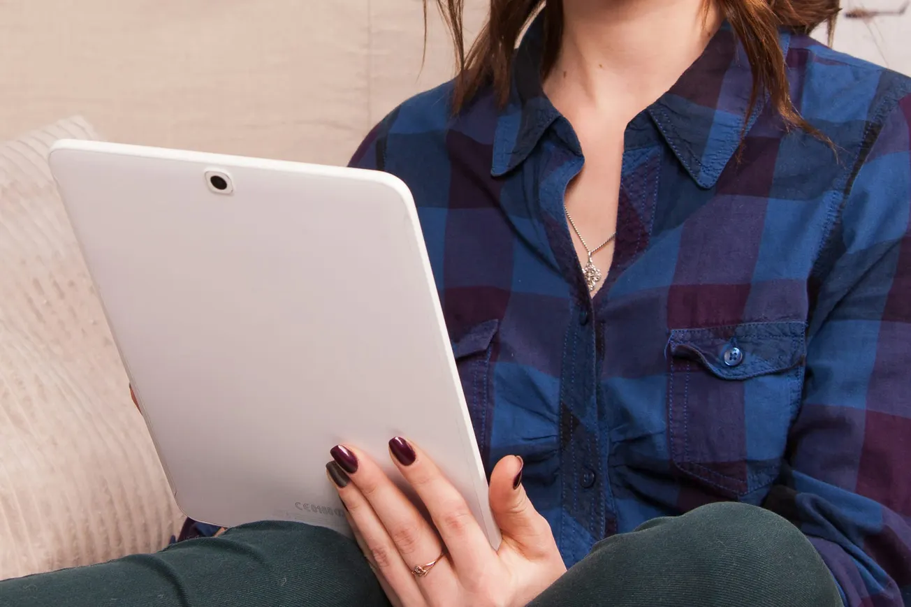 A person in a blue plaid shirt holds a white tablet, sitting on a striped cushion. The scene conveys a relaxed, casual atmosphere.