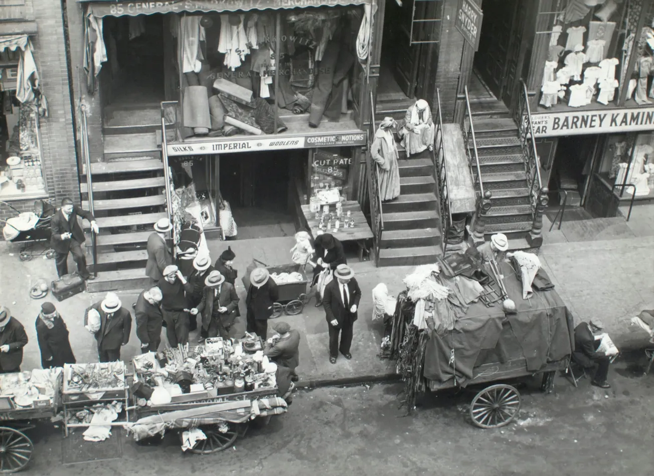 A bustling 1930s street market scene with vendors selling goods from carts, surrounded by formally dressed men in hats. Shops in the background display clothing.