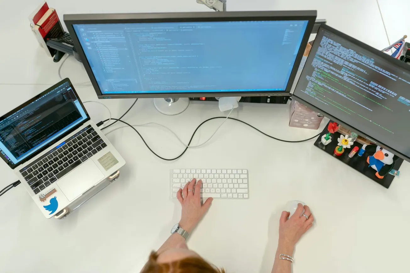 Overhead view of a person typing on a keyboard, surrounded by two large monitors and a laptop displaying code, suggesting a focused work environment.