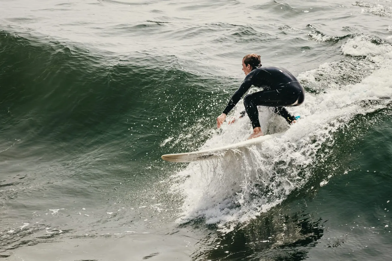 A surfer in a black wetsuit rides a large, curling wave on a white surfboard. The water splashes dynamically, conveying excitement and motion.