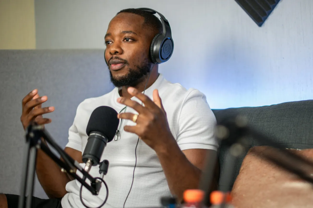 A man wearing headphones speaks passionately into a microphone, seated in a cozy studio with acoustic foam panels. He gestures expressively, conveying engagement.