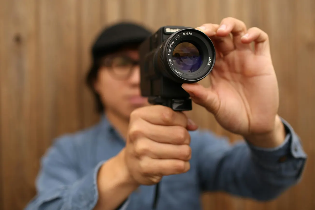 A person wearing a blue shirt and black cap holds a vintage video camera, focusing the lens. The background is a wooden wall, creating a nostalgic atmosphere.