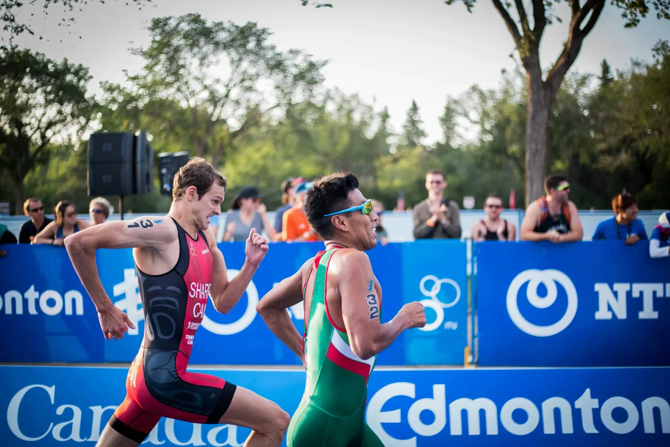 Two triathletes sprint side by side during a race, surrounded by cheering spectators. Trees and event signage are visible in the background.