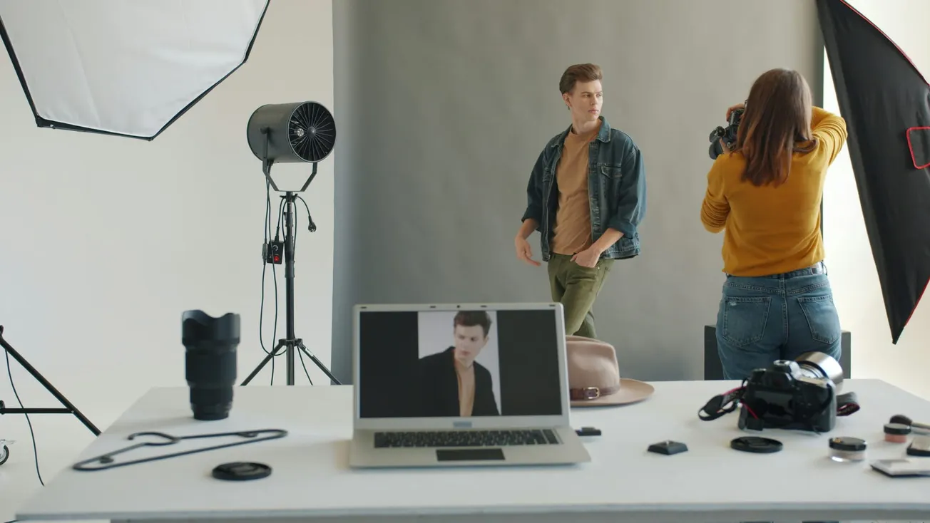 A photographer in a yellow sweater photographs a model in a denim jacket in a studio. A laptop displays a portrait, and camera gear is on the table.